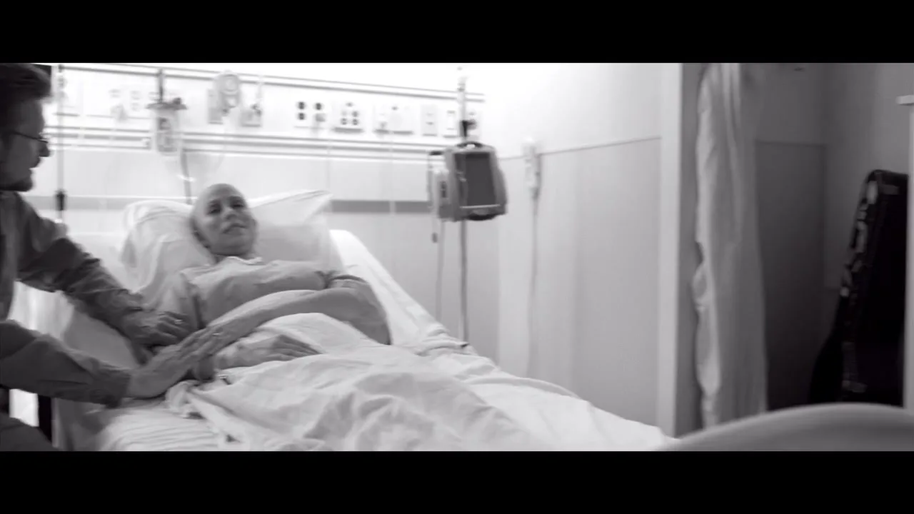 A man comforts a woman in a hospital bed, then a woman with a headscarf helps a girl with homework at a desk by a window.