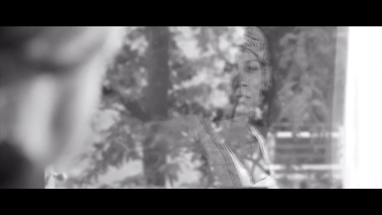 A woman with a headscarf looks out a window, her reflection visible, in a black and white shot.