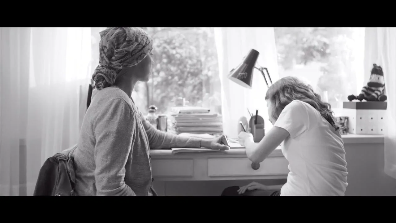 A woman with a headscarf sits at a desk next to a young girl who is writing, both looking out a window in a bright room.