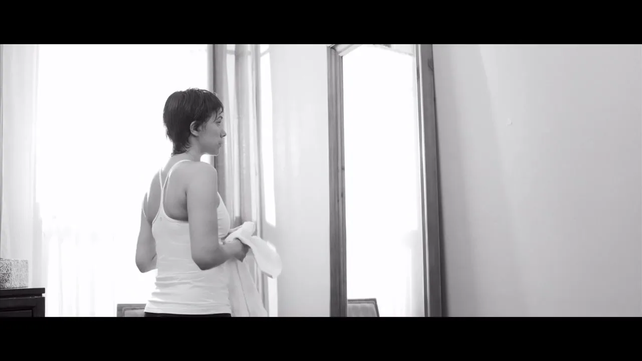A woman with short hair, wearing a tank top, stands in a room, looking out a window, in black and white.