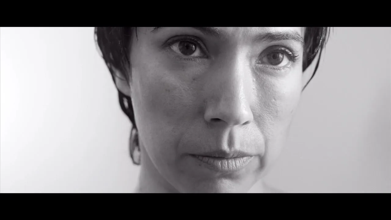 A woman with short dark hair looks directly at the camera with a serious expression in a black and white medium closeup shot.