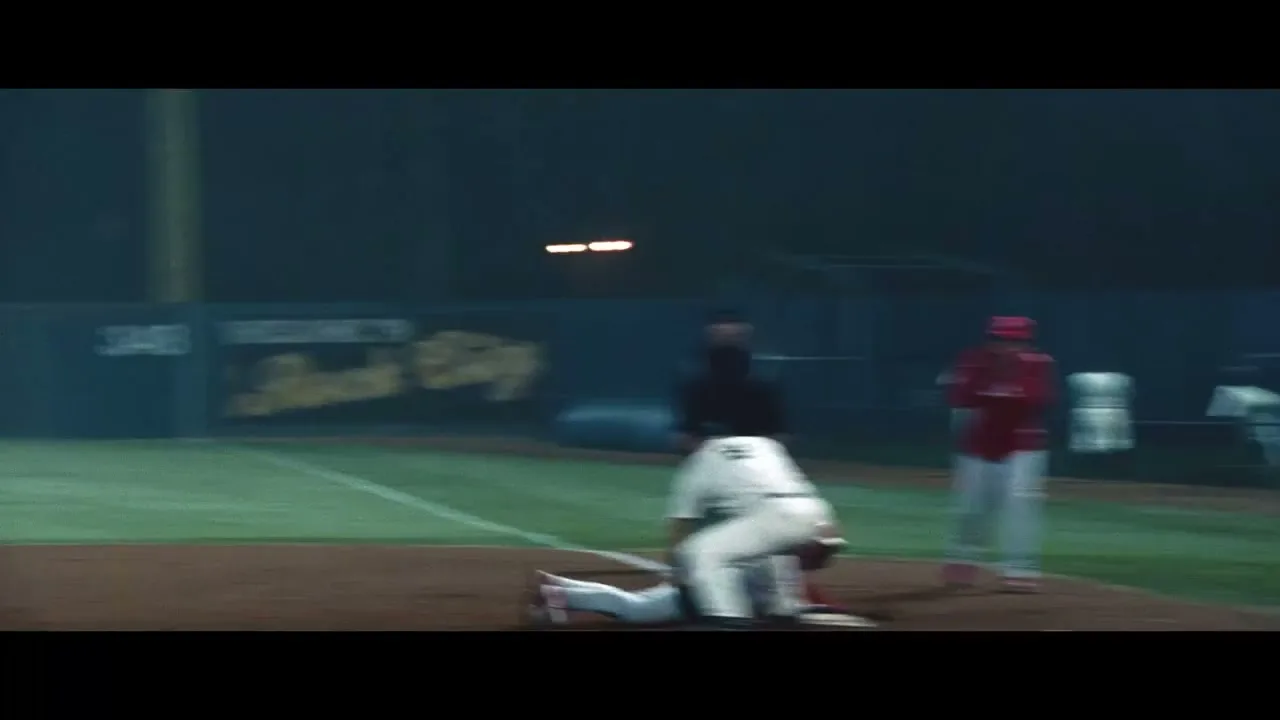 A baseball player in a white uniform and black cap stands on a baseball field at night, then slides into a base, and then stands up again.