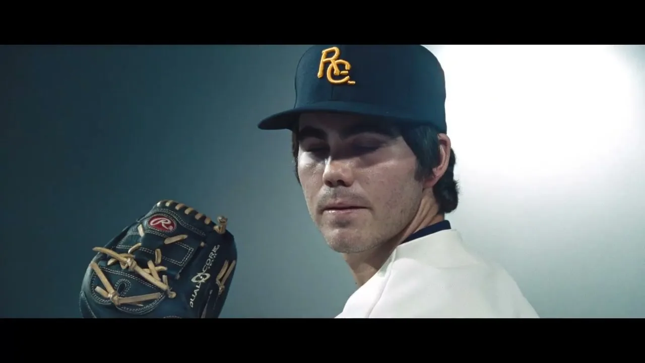 A male baseball player in a navy blue cap and white jersey looks intently at the camera, holding a Rawlings glove in his left hand.