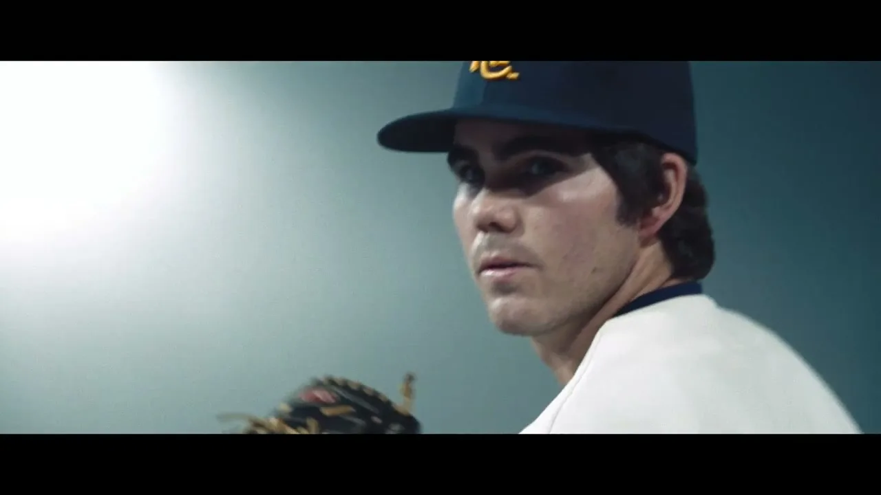 A young male baseball player in a navy blue cap and white jersey looks intently at the camera, holding a baseball glove.