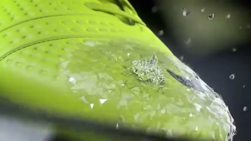 A close-up of a bright green Nike soccer shoe as water droplets hit its surface, creating a shimmering effect.