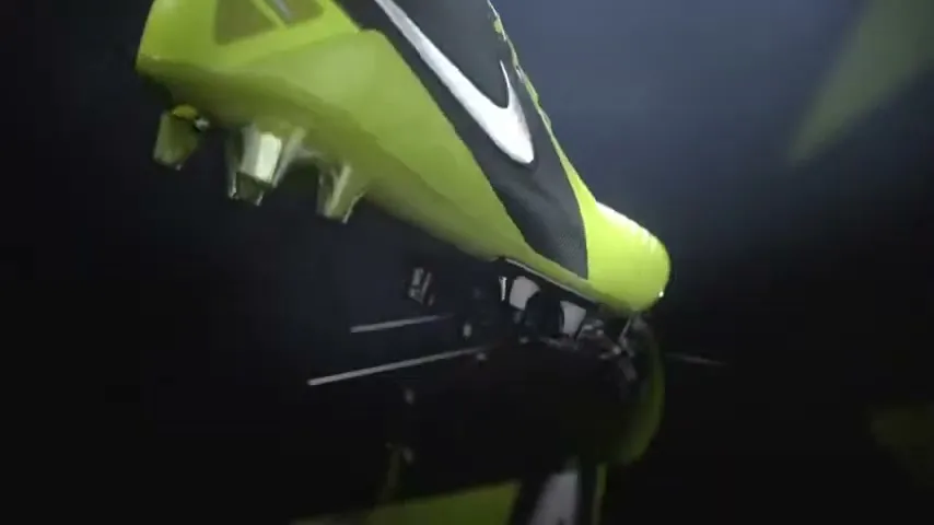 A bright green and black Nike soccer cleat with a white swoosh logo is shown in a studio, with small debris flying off its sole.