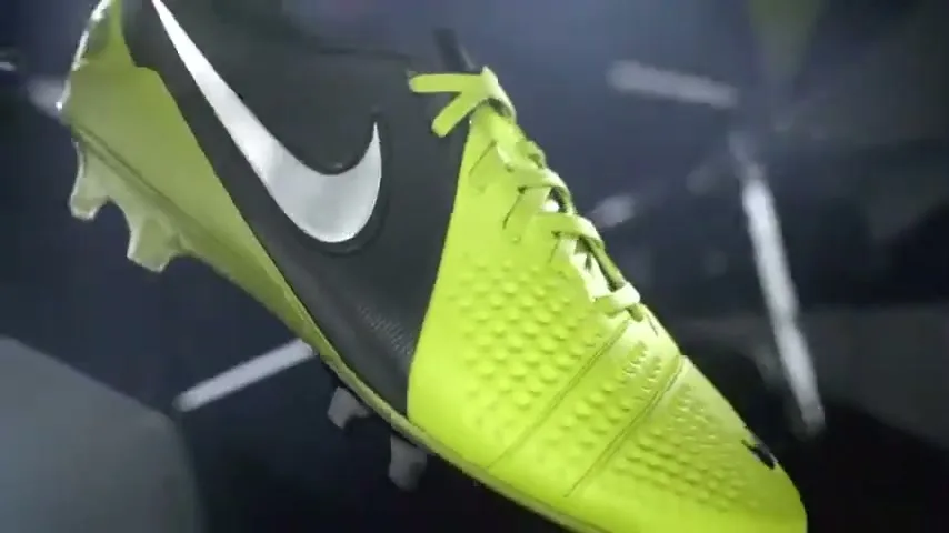 A neon green and black Nike soccer cleat rotates in a dark, abstract space with shards of light.