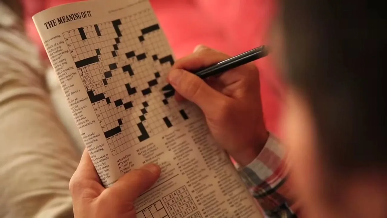 A person's hands hold a newspaper crossword puzzle, filling in answers with a pen, in a brightly lit interior setting.