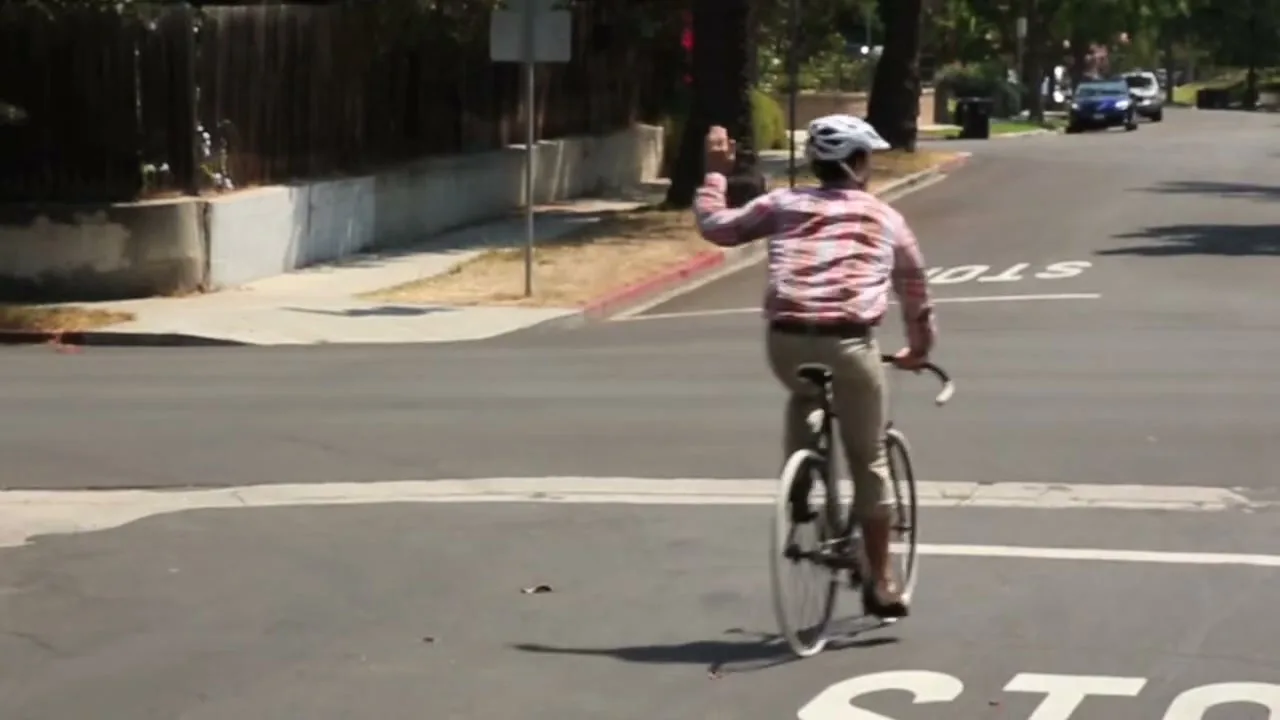 A man in a plaid shirt and helmet rides a bicycle, stopping at a stop sign on a sunny suburban street.