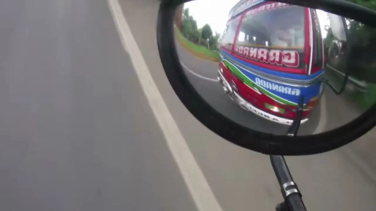 A colorful bus is reflected in a bicycle's rearview mirror, driving on a road with trees in the background.