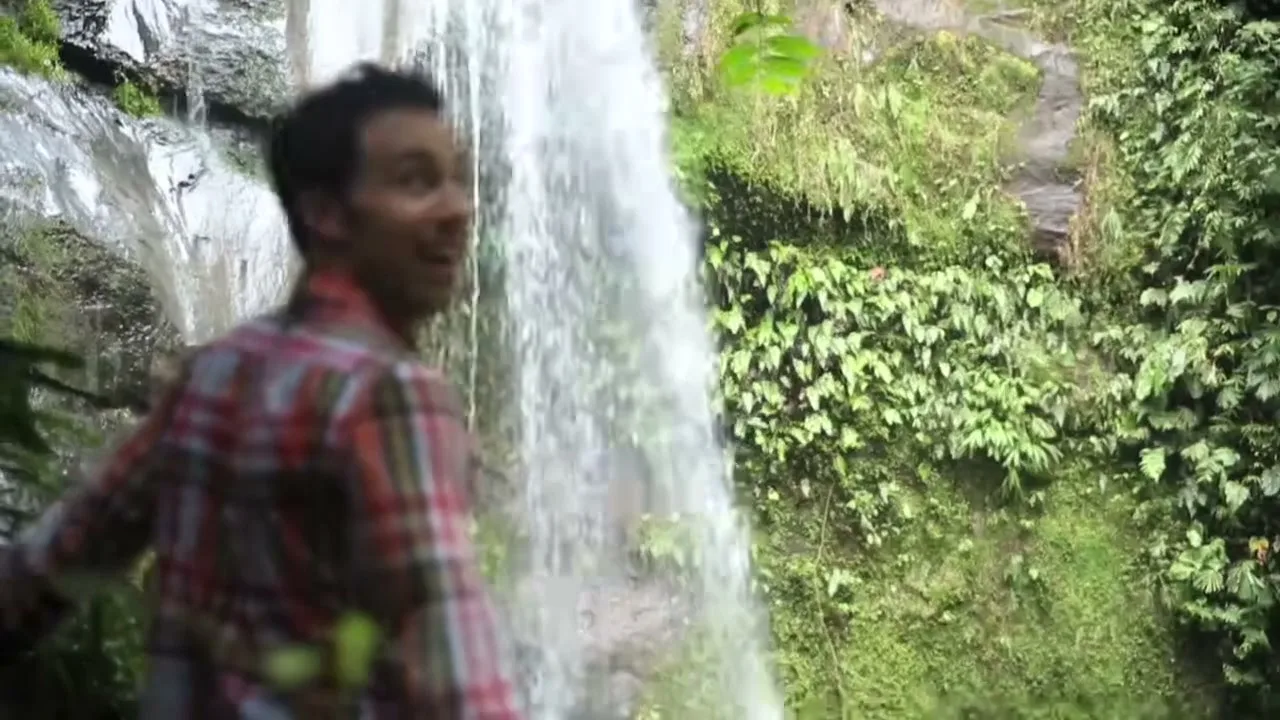 A man in a plaid shirt stands next to a waterfall, looking surprised and happy in a lush green jungle.