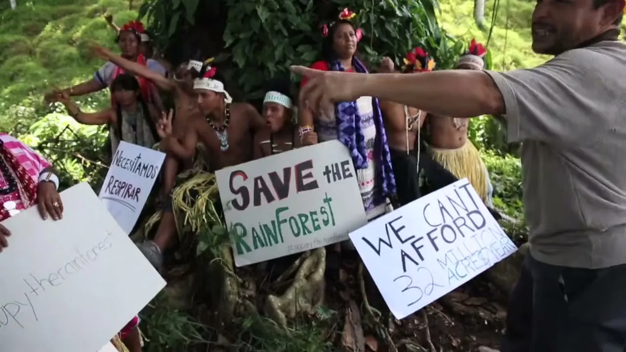 A group of indigenous people and a construction worker protest deforestation in a lush rainforest, holding signs that read "SAVE THE RAINFOREST" and "WE CAN'T AFFORD 32 MILLION ACRES."