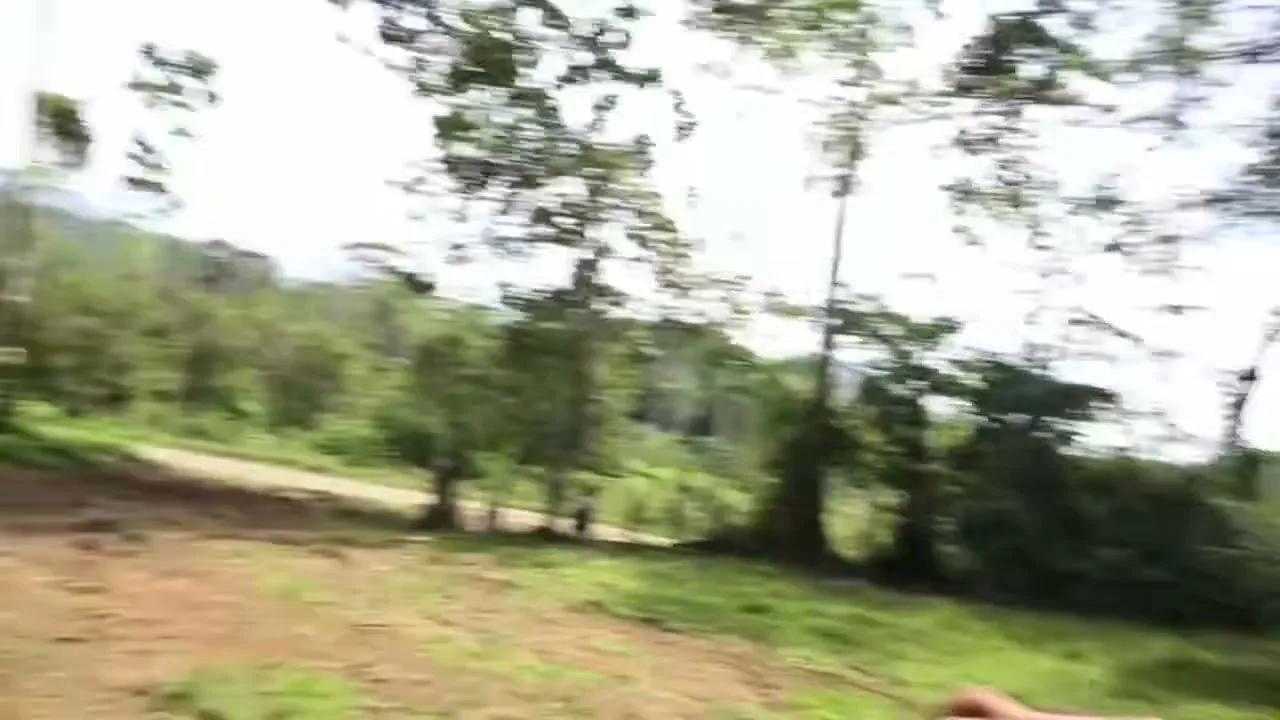 A man with blue face paint and a man in an orange shirt are seen with other workers in a rural, outdoor setting, with trees and hills in the background.