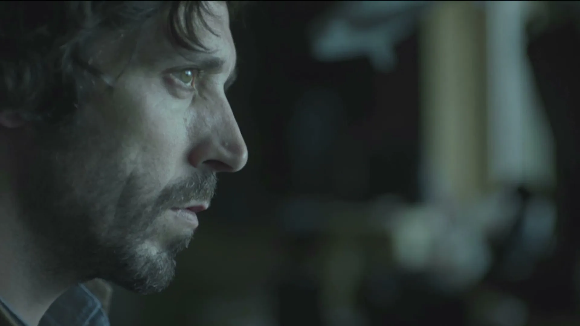 A man with a beard and dark, curly hair looks intently off-screen, his face illuminated by an unseen light source in a dark room.