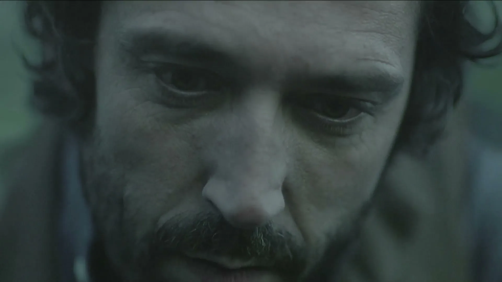 A man with dark, curly hair and a beard looks down, then slowly raises his eyes to look forward, in a close-up shot.