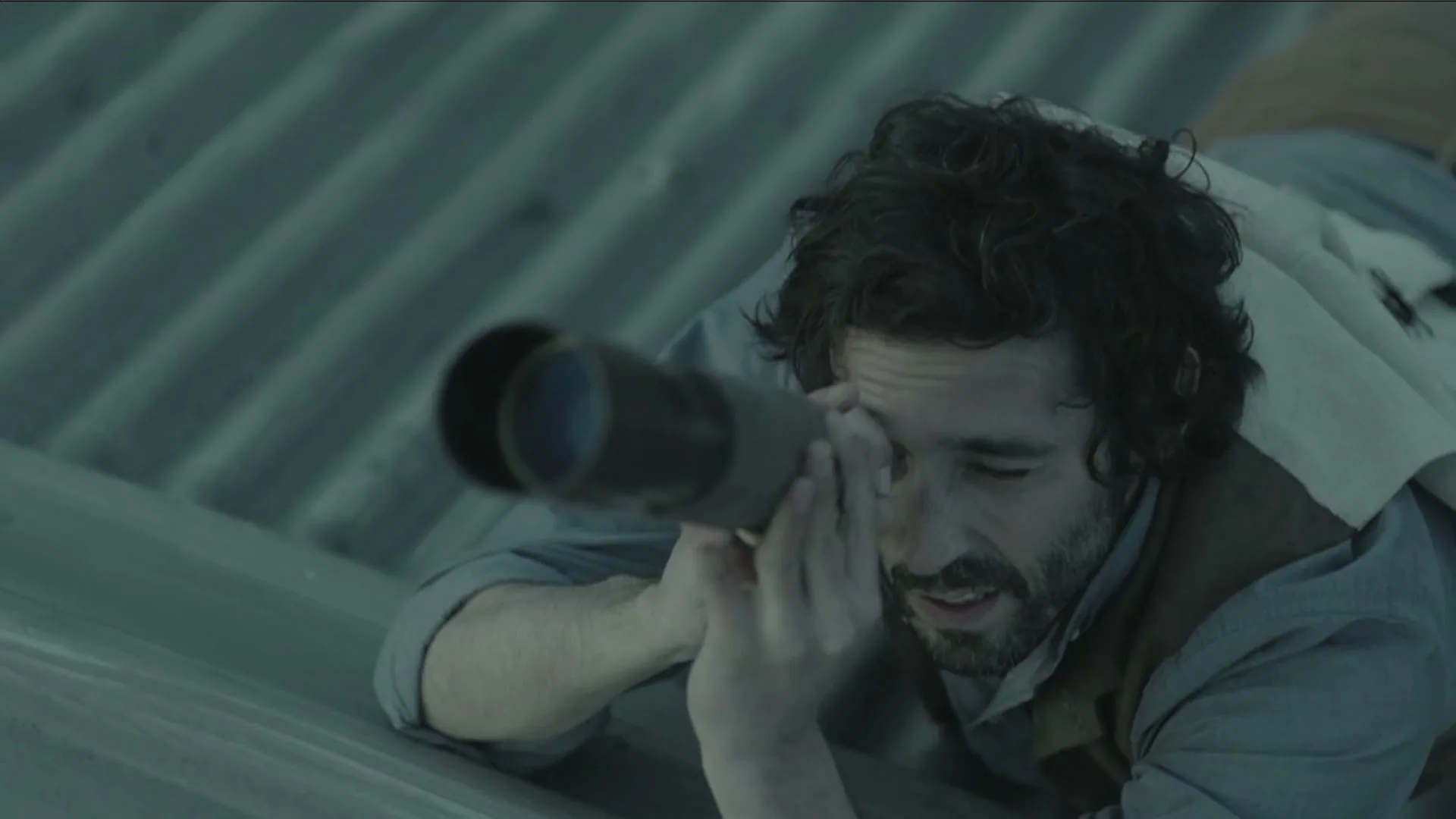 A man with dark curly hair and a beard lies on a corrugated metal roof, looking through a telescope.