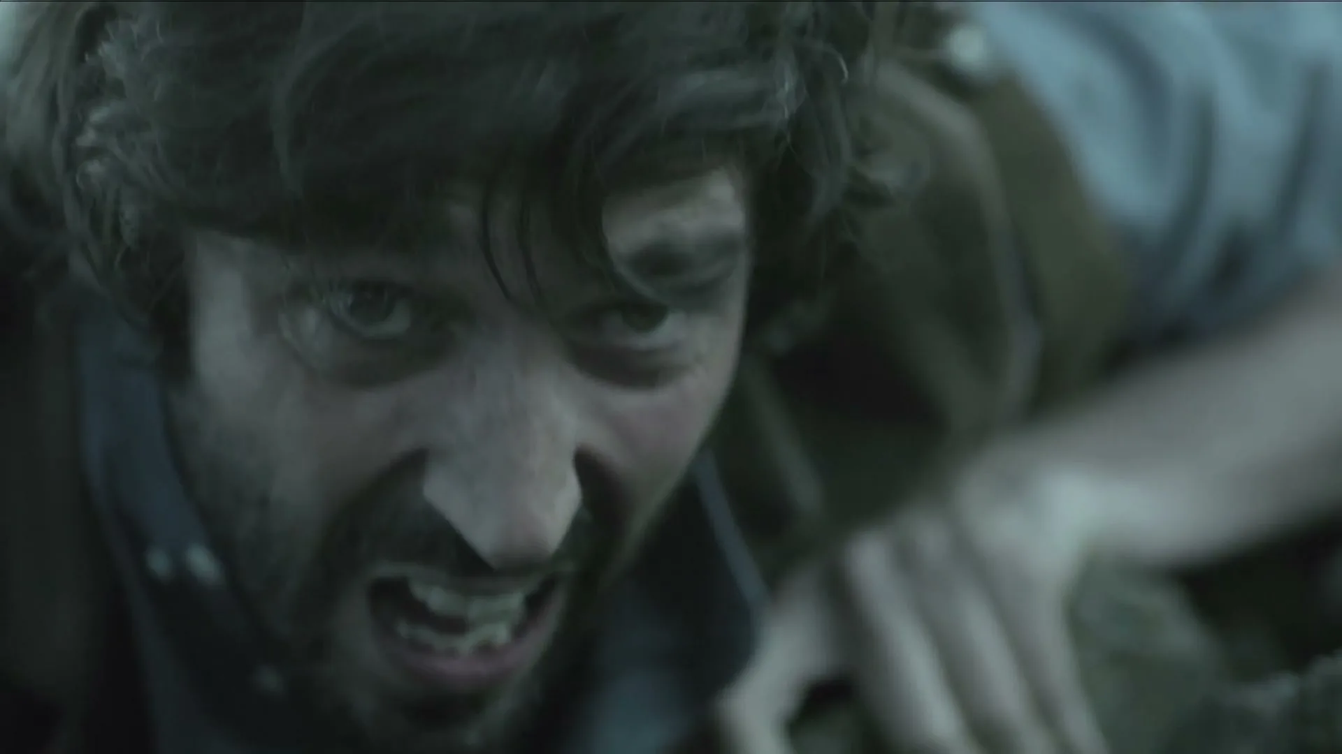 A man with a beard and messy hair struggles to climb, his face contorted in effort, as he reaches for a rock.