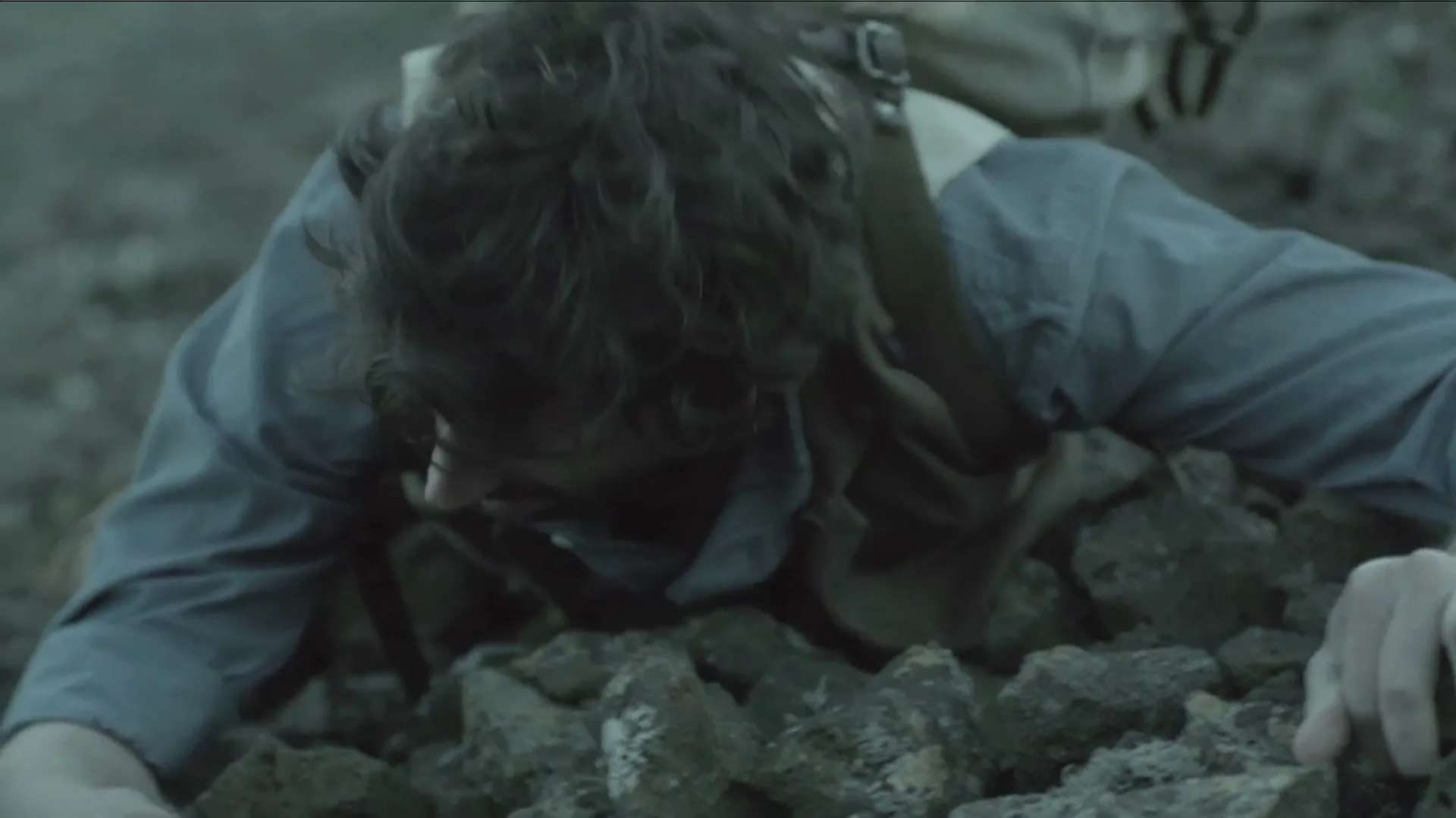 A man with dark, curly hair and a beard, wearing a blue shirt and a brown vest, struggles to crawl over a pile of rocks.
