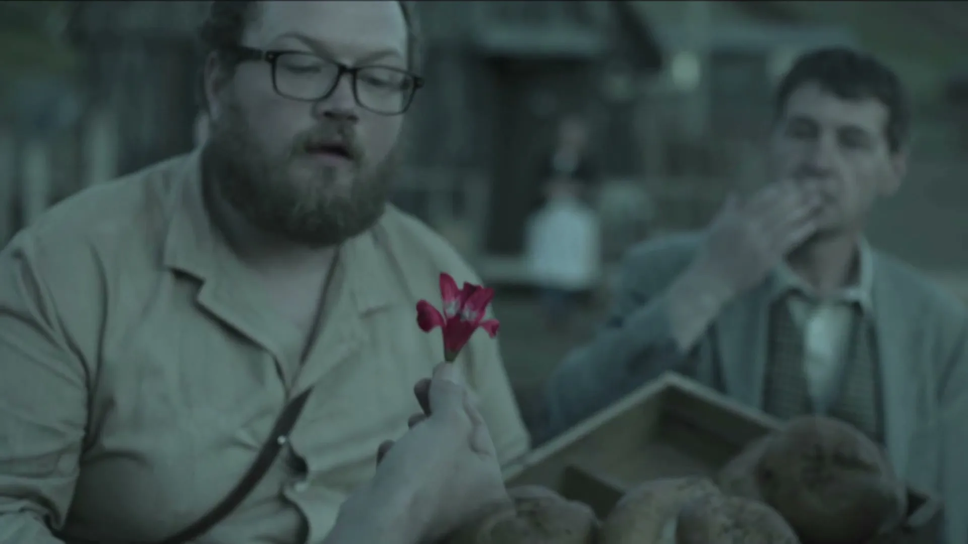 A bearded man in glasses looks at a small red flower held out to him, while another man watches in the background.