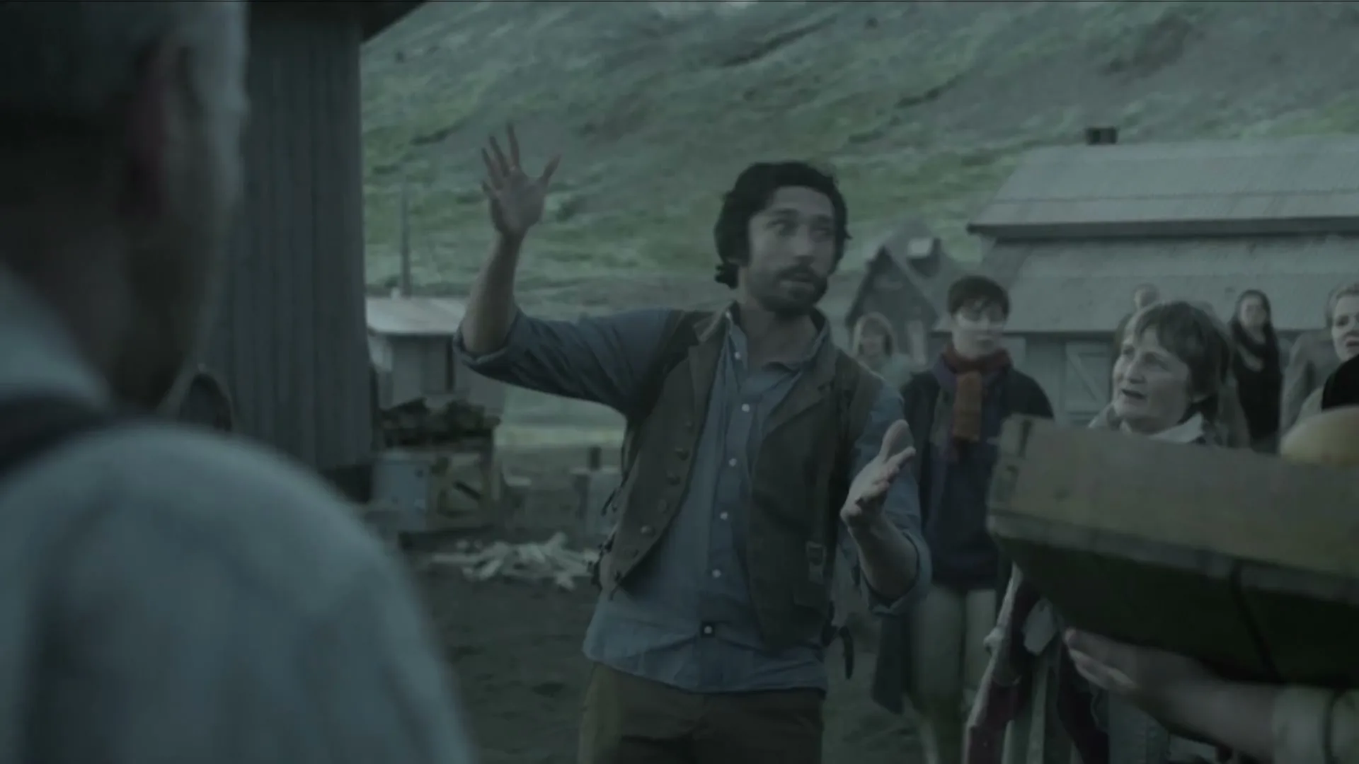 A man with dark hair and a beard gestures with his hands while talking to an unseen person, surrounded by other people in a desolate, rural village.