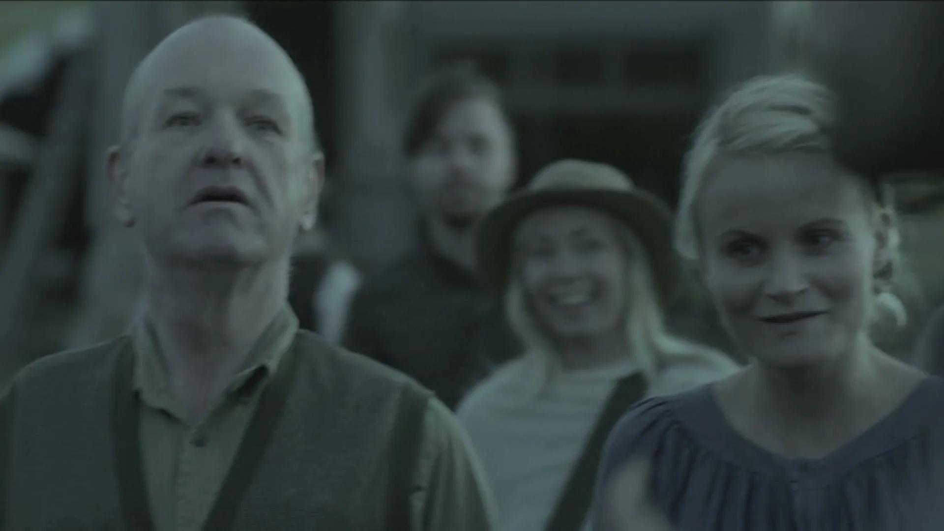 A diverse group of people, including a blonde woman and a bald man, stand together outdoors, looking on with expressions of hope and anticipation.