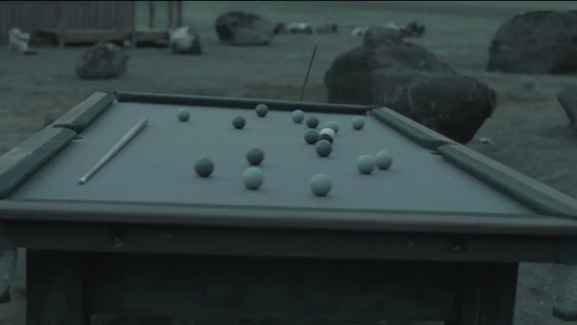 A wide shot of a pool table with grey and white balls scattered on its surface, outdoors in a desolate, rocky landscape.