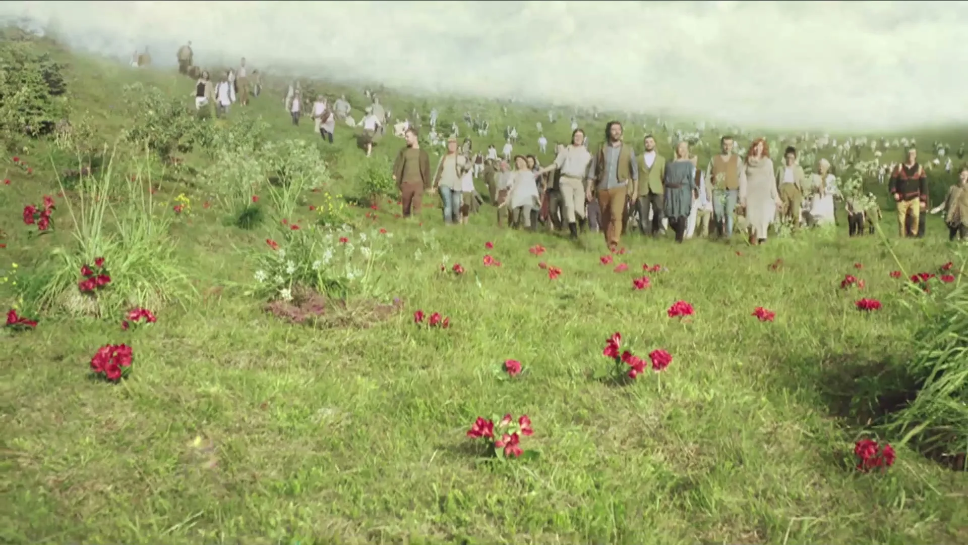 A diverse group of people, dressed in earthy tones, walk together up a grassy hill dotted with red flowers under a bright, overcast sky.