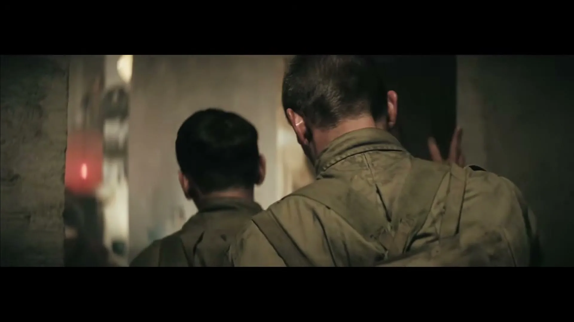 Two men in military-style uniforms walk through a dark, gritty corridor, looking over their shoulders at a mysterious figure in the background.