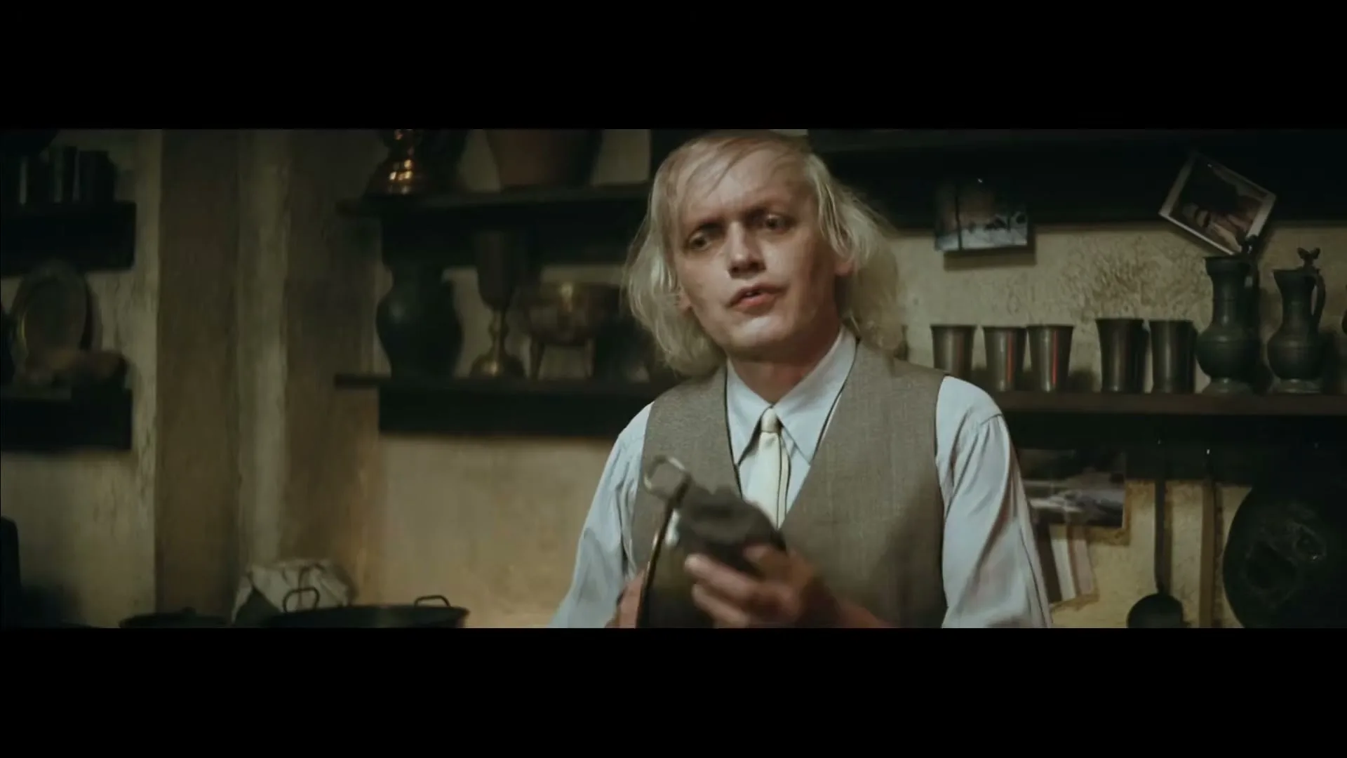 A pale man with blonde hair and a vest holds a small object in a rustic kitchen, looking directly at the camera.