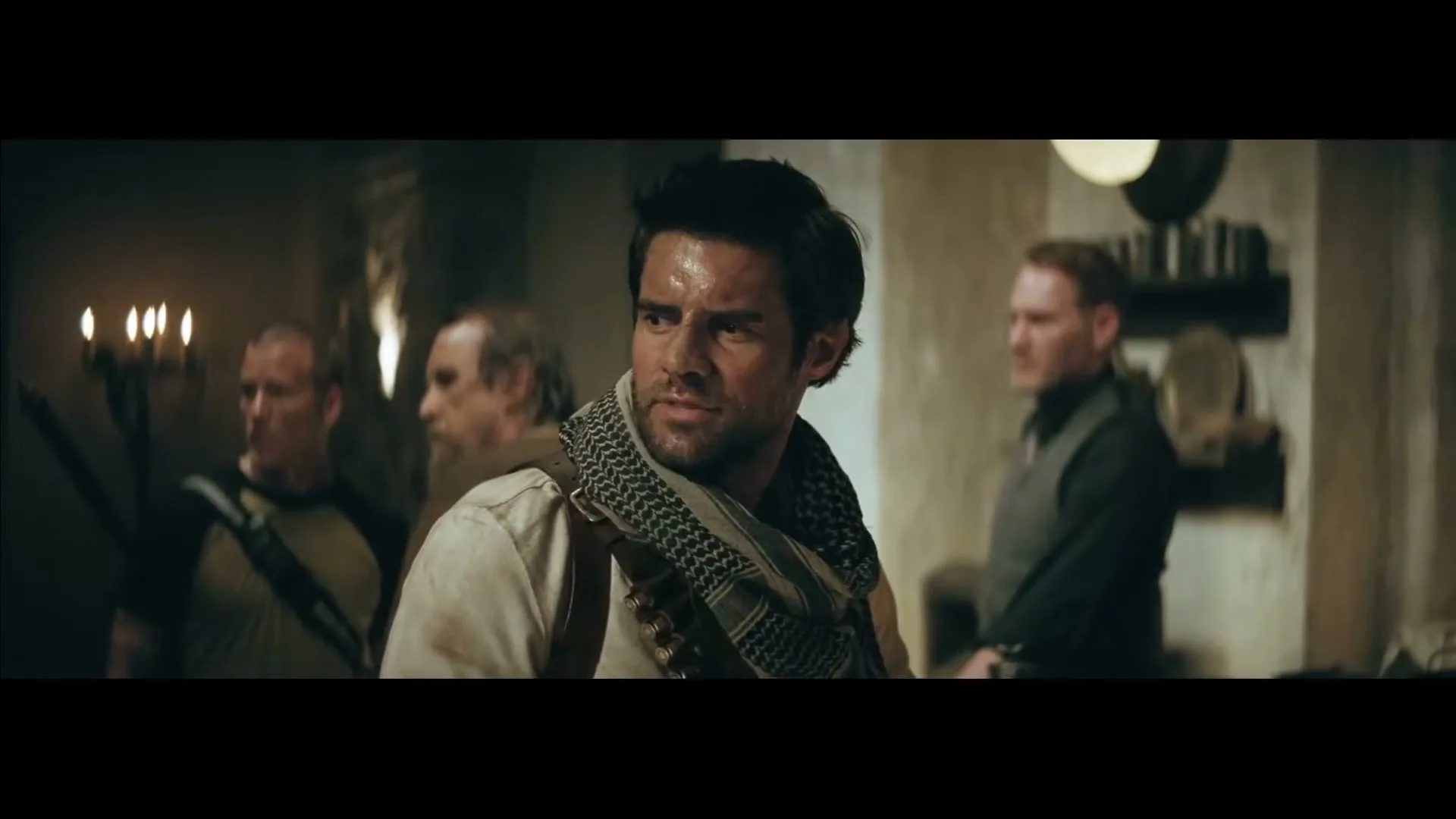 A rugged man with a scarf and bandolier looks intently to the side, surrounded by other men in a dimly lit, rustic interior.