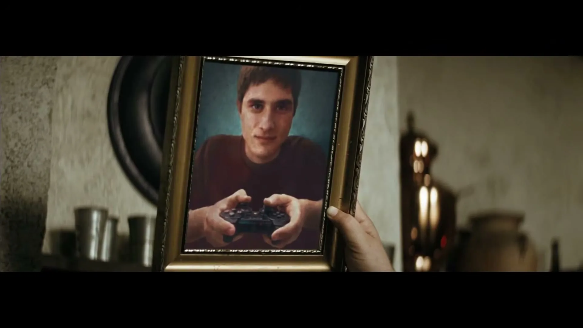 A person's hand holds up a framed photo of a young man with short brown hair, wearing a maroon shirt, holding a PlayStation controller, and smiling slightly.
