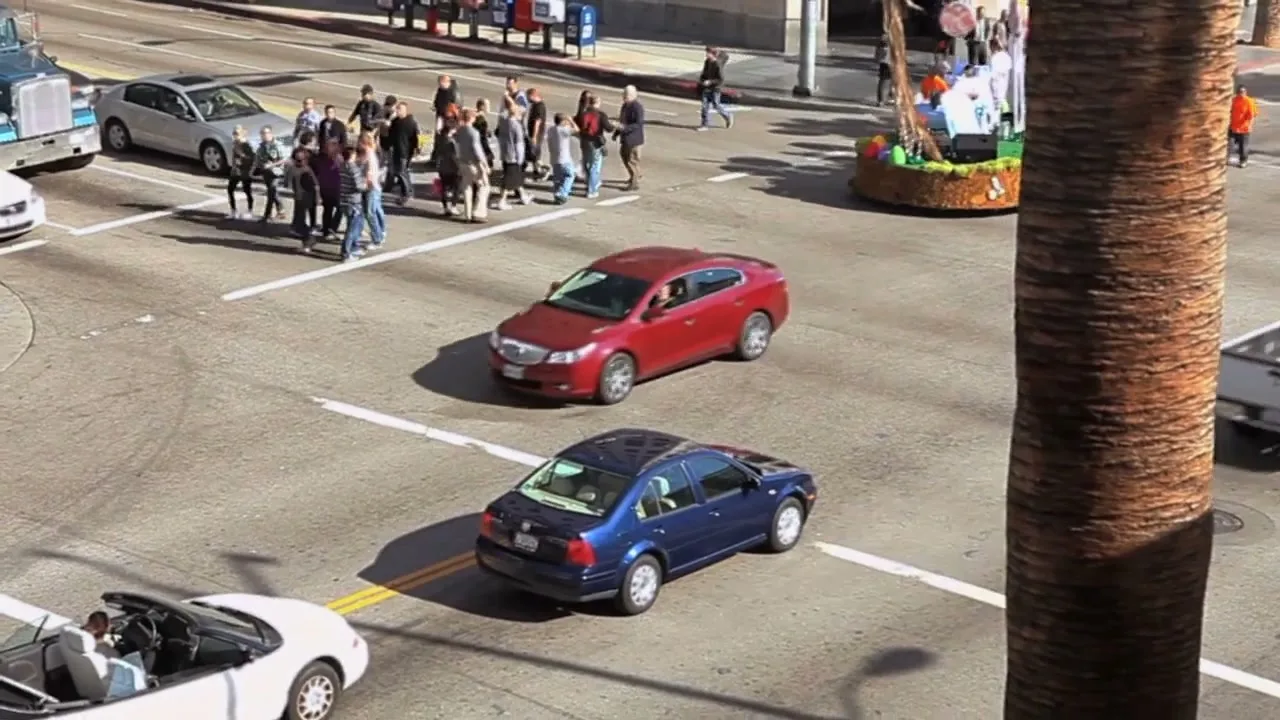 A high-angle shot captures a busy street intersection with cars and pedestrians, as a parade float passes by on the right.
