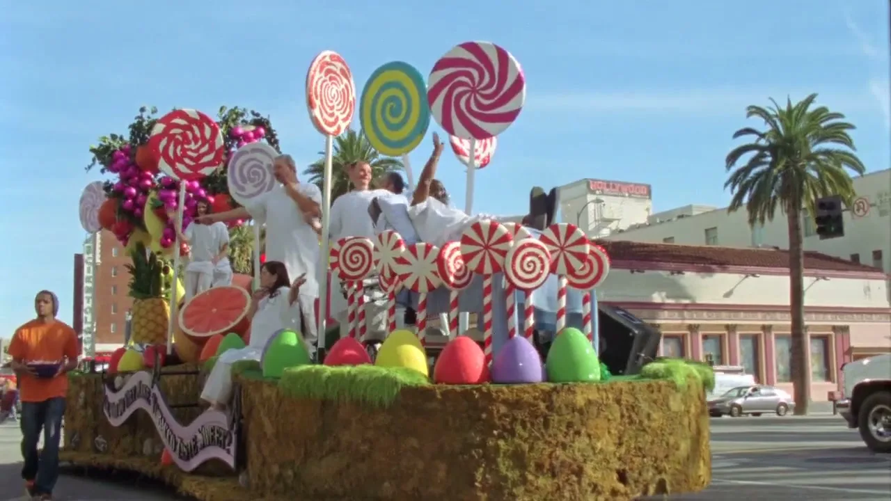 A parade float resembling a giant cake with lollipops and fruit moves down a sunny street, with people in white outfits on top and a man walking alongside.