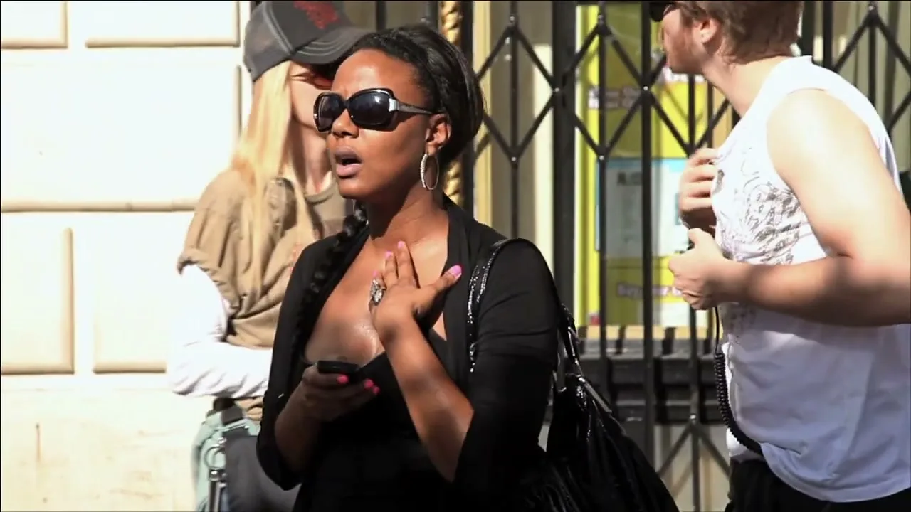 A young Black woman in sunglasses and a black top holds a phone and looks surprised, standing on a sunny city street with other people.