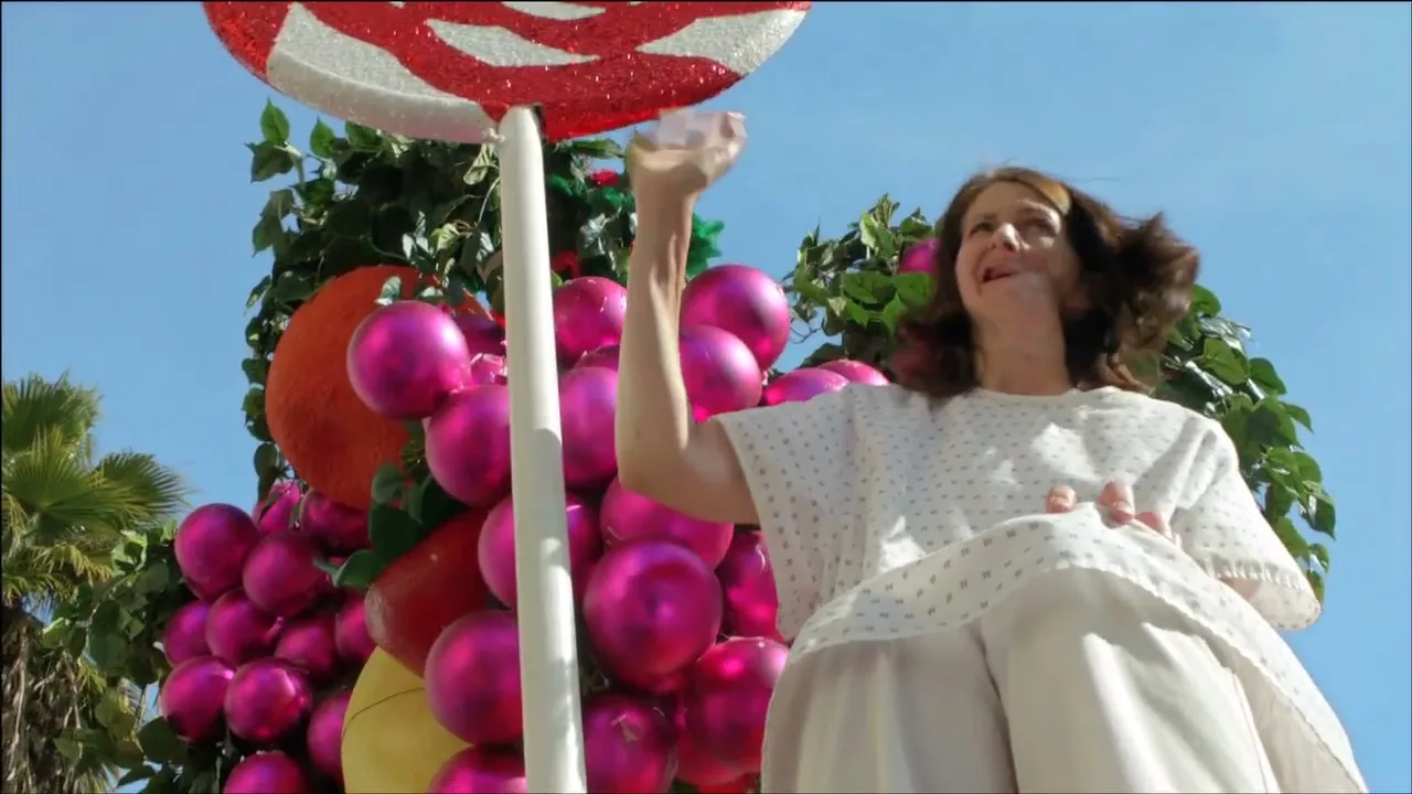 A woman in a hospital gown sits on a parade float decorated with oversized fruit and a lollipop, looking up and waving.