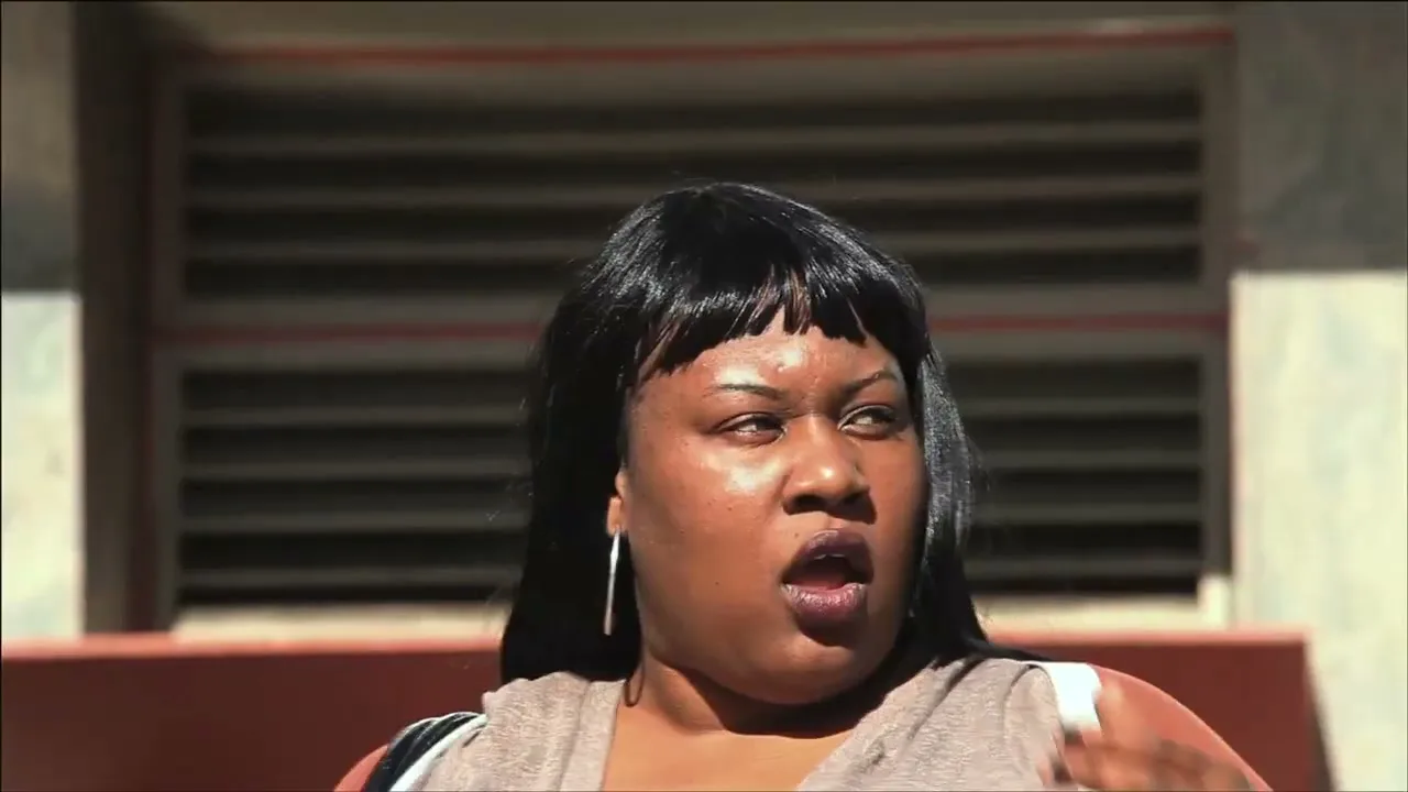 A Black woman with long black hair and hoop earrings looks surprised, mouth open, in an outdoor setting with a building in the background.