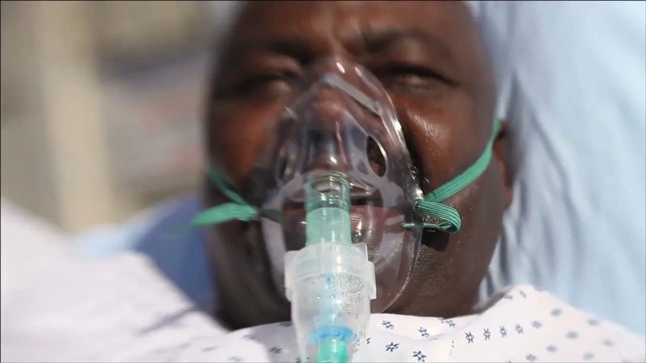 A close-up shot shows an older Black person lying in a hospital bed, wearing an oxygen mask, with their eyes closed.
