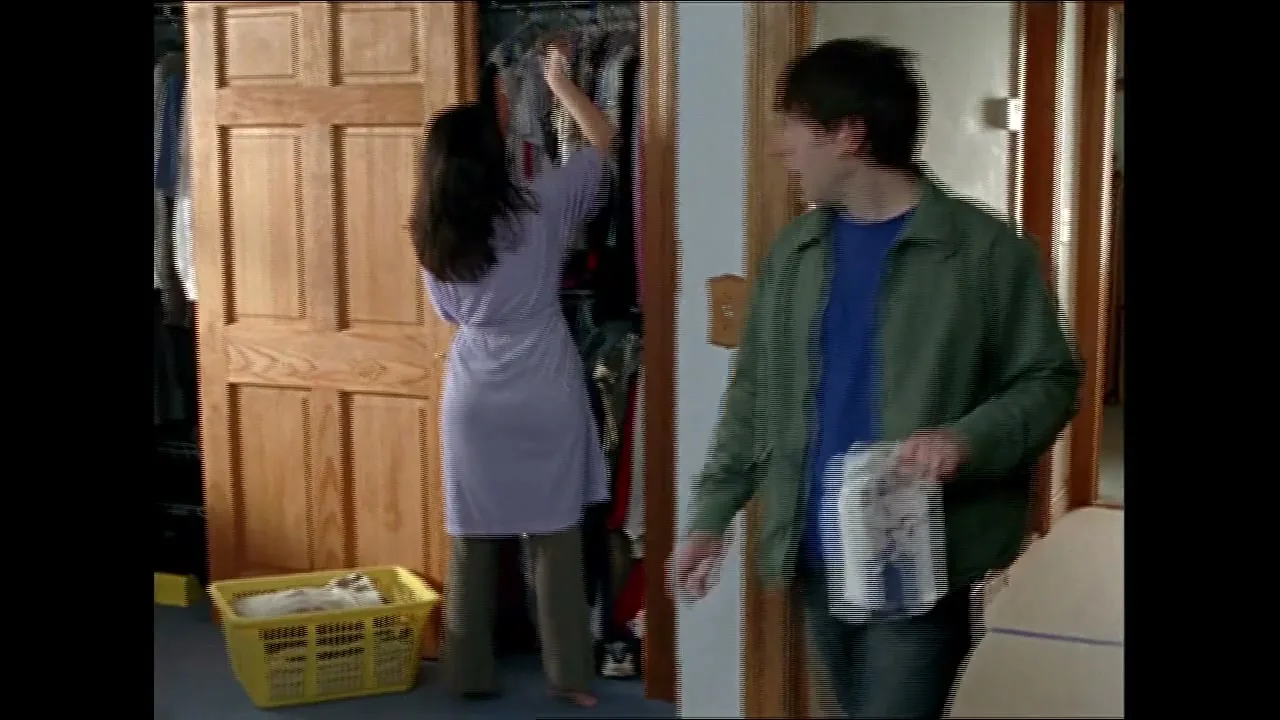 A woman in a purple robe looks through a closet while a man in a green jacket walks past her holding a box of eggs in a bedroom.