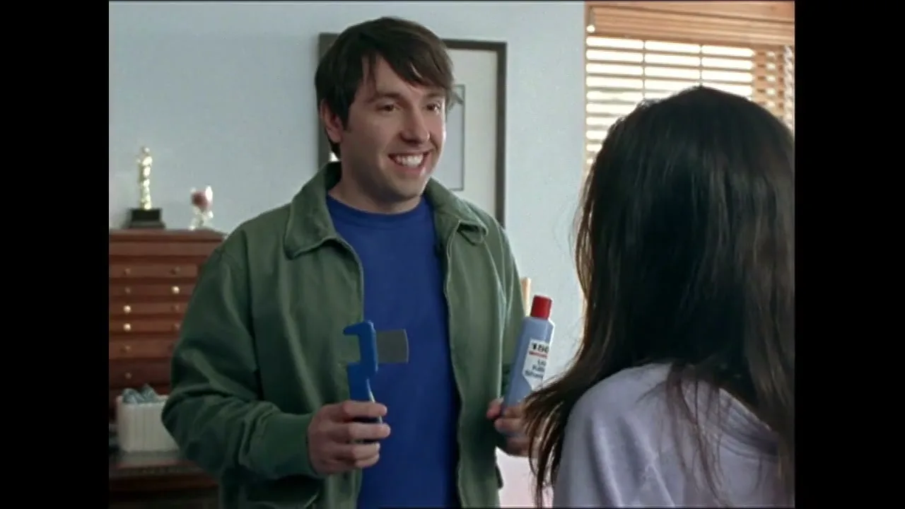 A man in a green jacket and blue shirt holds a blue wrench and a bottle of lice shampoo, smiling at a woman with long dark hair in a bright room.