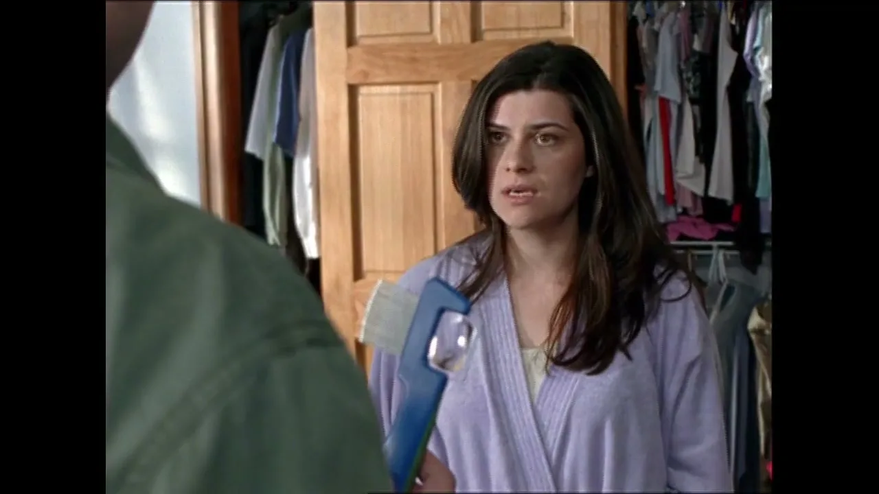 A woman in her 30s, wearing a purple robe, looks concerned while holding a blue lice comb in a bedroom closet.