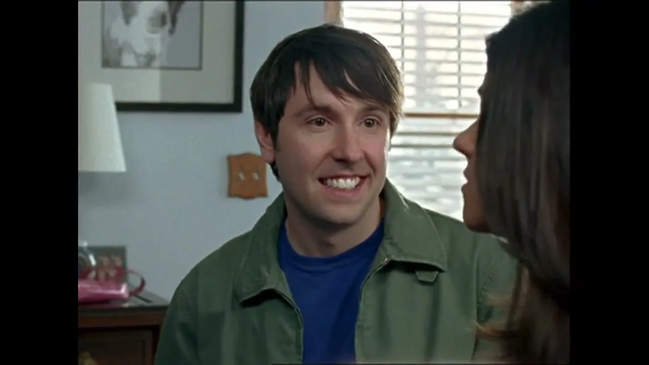 A man with dark hair and a green jacket smiles and talks to a woman with dark hair, who is partially visible, in a brightly lit room.