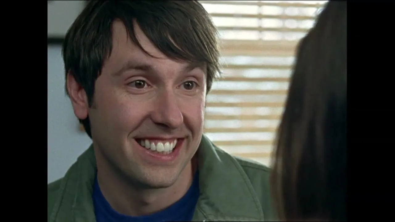 A man with dark hair and a green jacket smiles broadly while looking at someone off-screen in an interior setting.