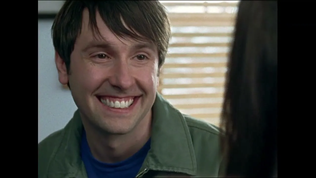 A man with dark hair and a green jacket smiles broadly, revealing white teeth, as he looks at someone off-screen in an interior setting.
