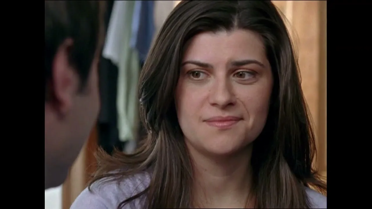 A woman with long dark hair looks at a man off-screen, her expression shifting from serious to a slight smile in an interior setting.