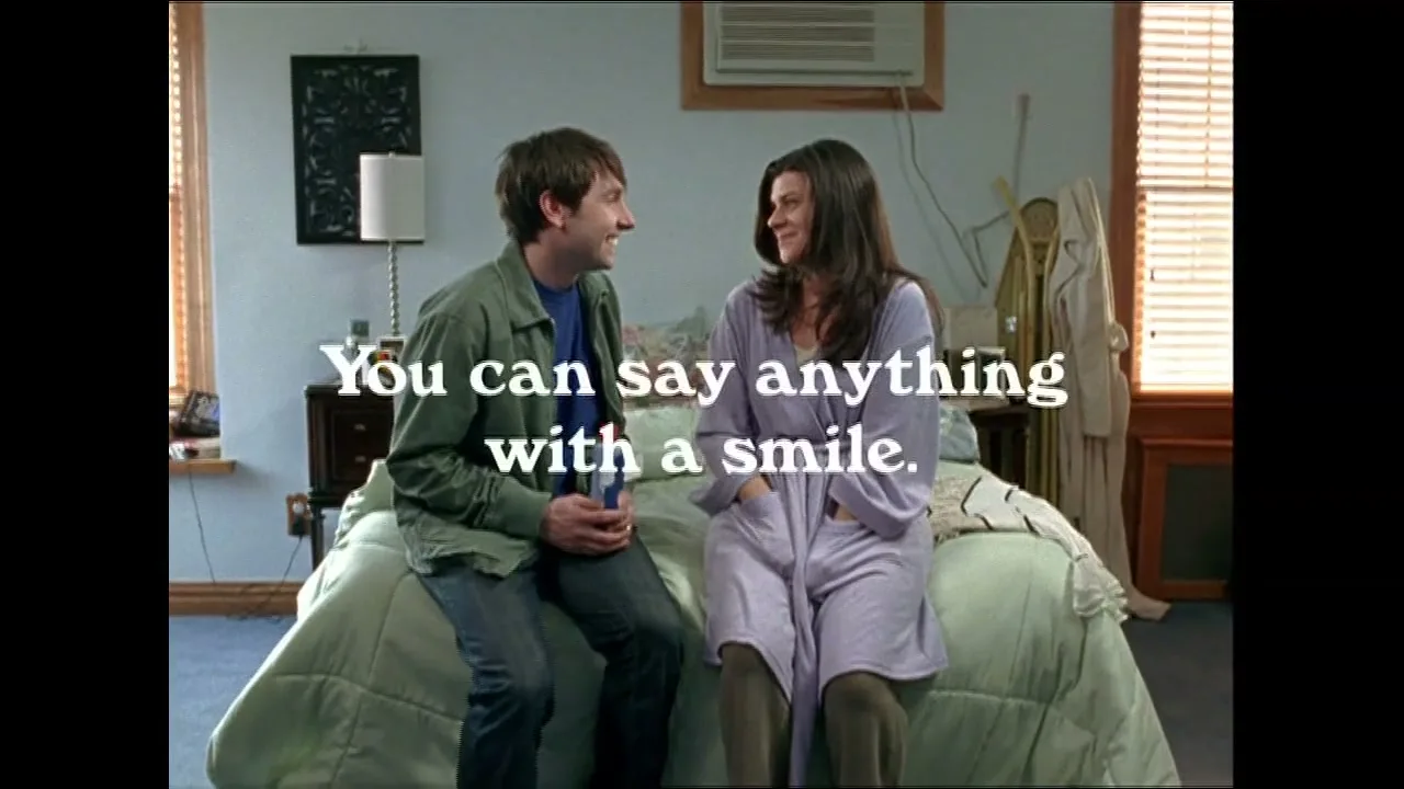 A man and a woman sit on a bed, smiling at each other, as the man holds a bottle of Crest mouthwash in a bedroom.