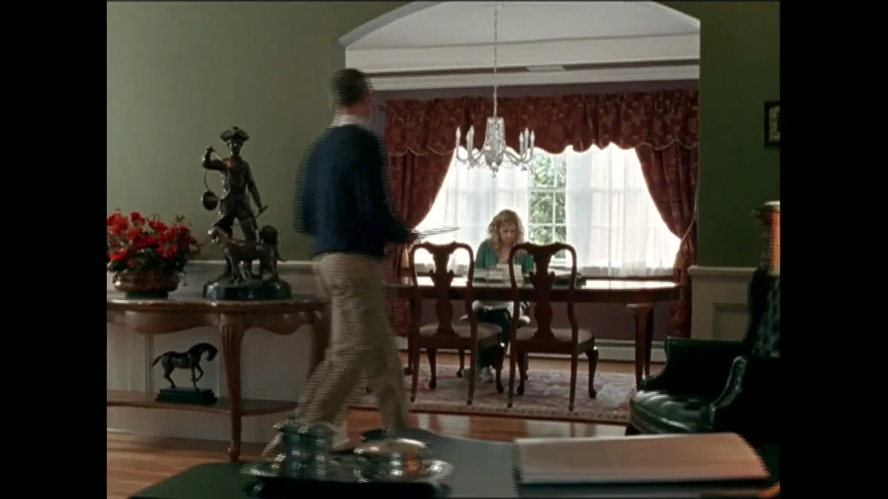 A man in a blue sweater and khaki pants walks past a table with a statue, towards a woman sitting at a dining table in a formal dining room.