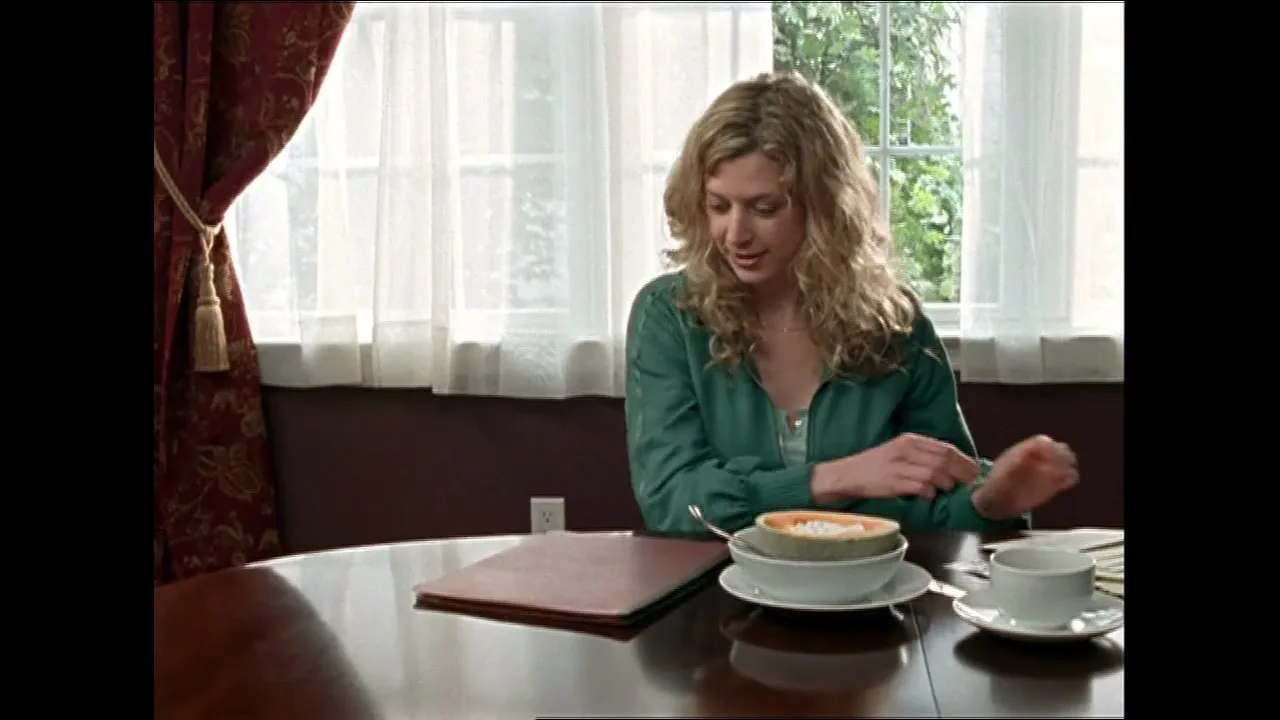 A blonde woman in a green shirt sits at a round table, eating fruit, as a man's arm places a document in front of her in a dining room.