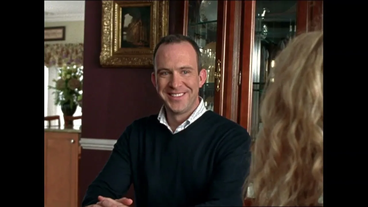 A man in his 30s, wearing a dark blue sweater and a collared shirt, smiles at a woman with blonde hair, sitting at a dining table in a home.
