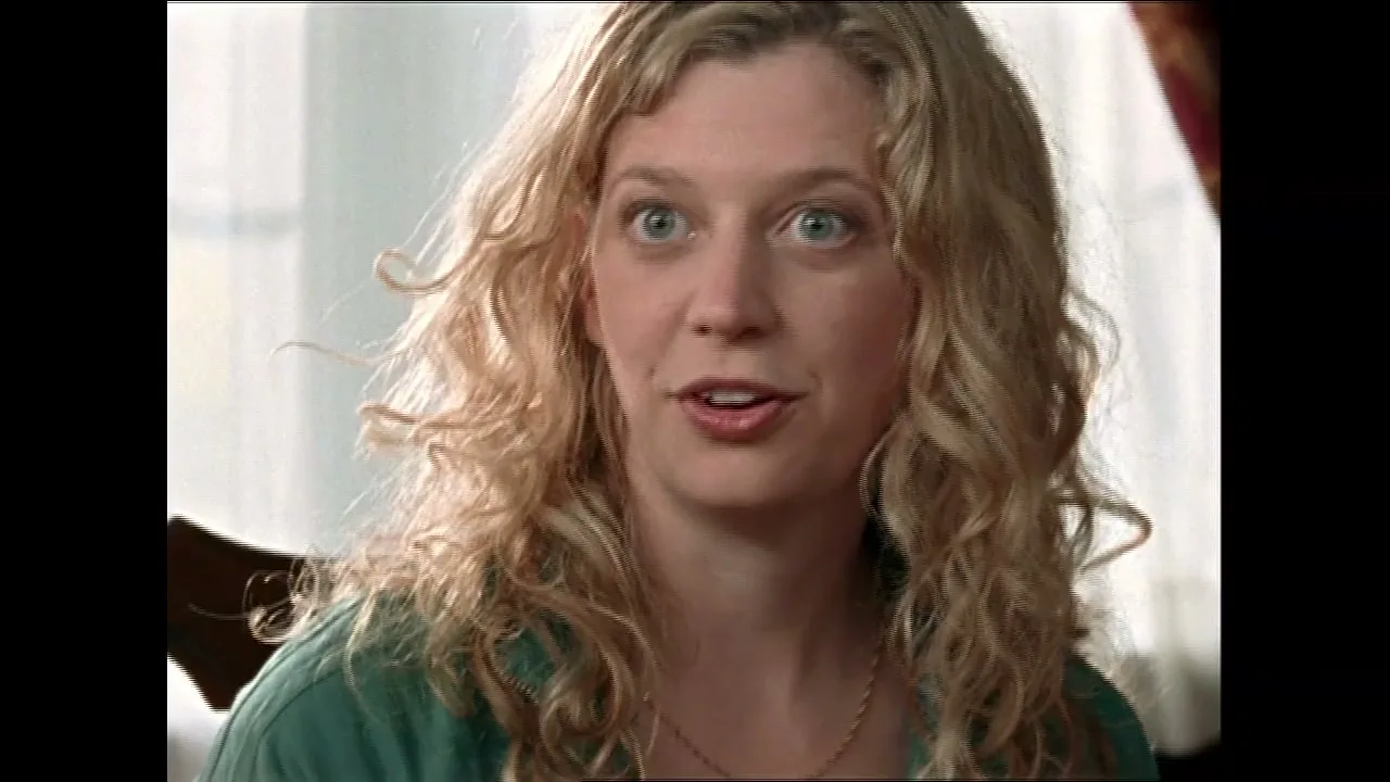 A blonde woman with curly hair and wide eyes looks off-camera, appearing surprised in an interior setting.