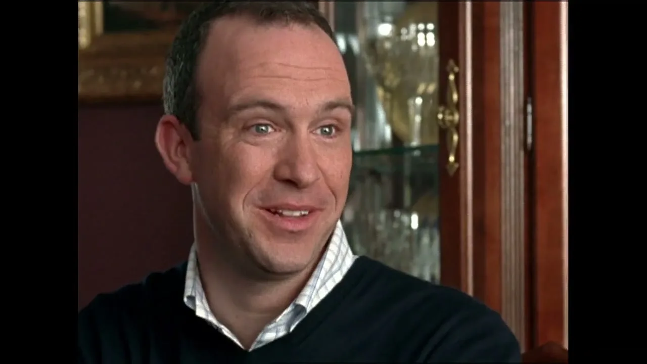A balding man in his 30s, wearing a dark sweater and collared shirt, smiles while looking off-camera in an interior setting.